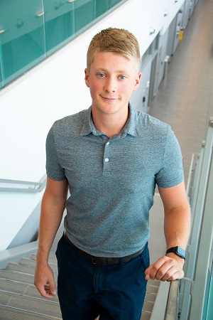 Young male student stands on staircase in M building at the Barrie Campus, wearing greenish/grey golf shirt and dark pants