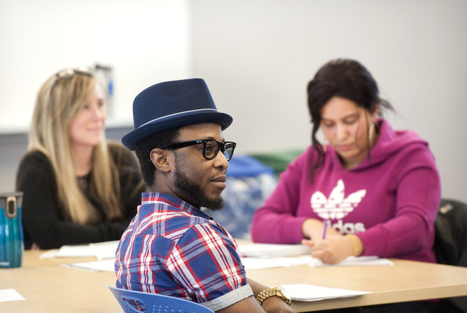 Student with hat on sitting at desk listening with 2 young women in the background
