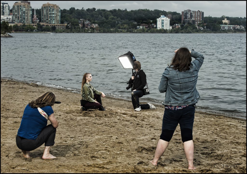 a female student kneeling and posing on the beach while three other students using lighting and camera equipment to capture her photo