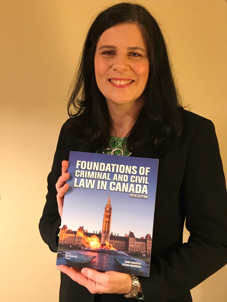 A person stands and smiles at the camera while holding a textbook reading "Foundations of Criminal and Civil Law in Canada, Fifth Edition."