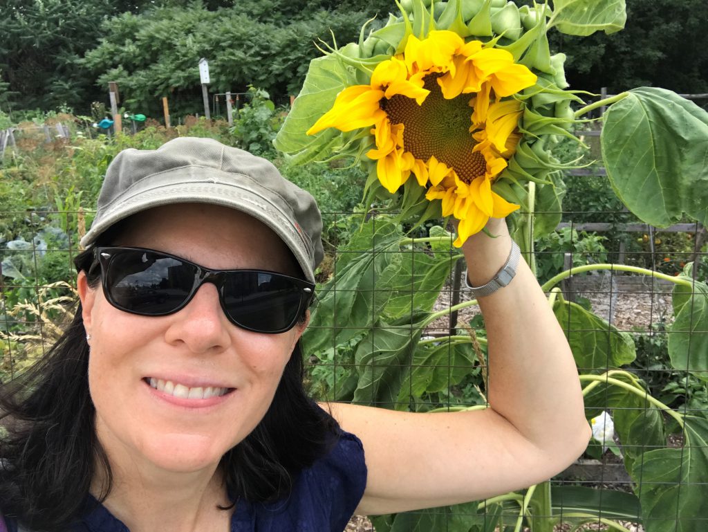 A person smiles at the camera while holding a sunflower that's taller than them in an outside garden.