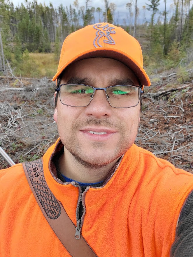 A person wearing glasses, orange vest and orange cap stands in the woods and smiles at the camera in a selfie.