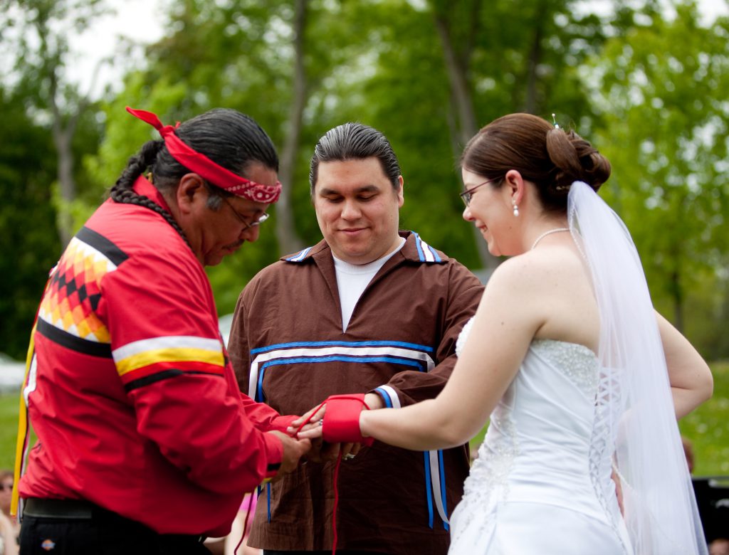 Two people, one in a wedding dress and the other in a brown shirt, stand outside with a third person in a red shirt who wraps their hands in red cloth.