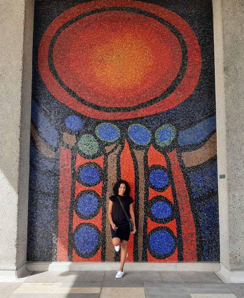 A person with black hair, black dress, white running shoes and black purse, smiles and leans against a red, blue, green and brown mosaic mural.
