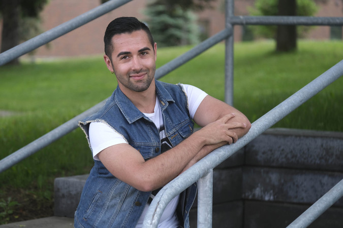 A person wearing a white T-shirt and blue jean jacket with no sleeves smiles and leans against a metal railing outside.