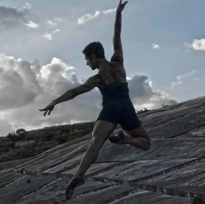 A dancer, pictured from behind, jumps across a rural landscape with one arm out to the side and the other held up above their head.