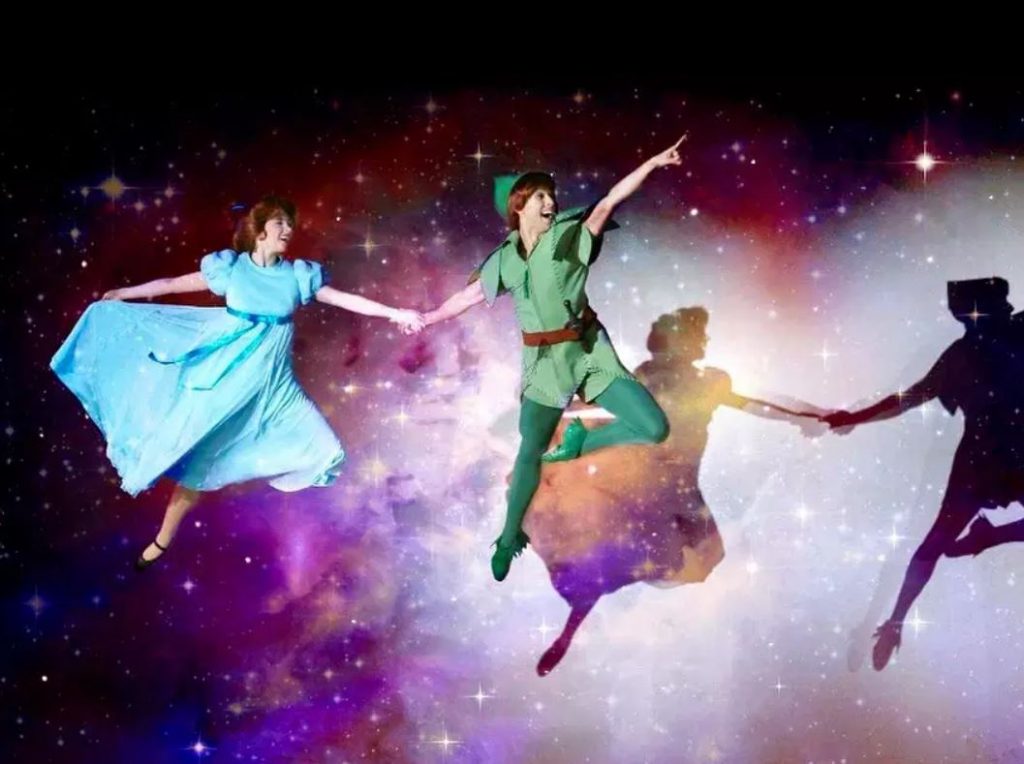 Two people dance through the air, held up by lines against a starry night background. They are dressed as Peter Pan in a green jumper, leggings and hat, and Wendy in a blue dress.