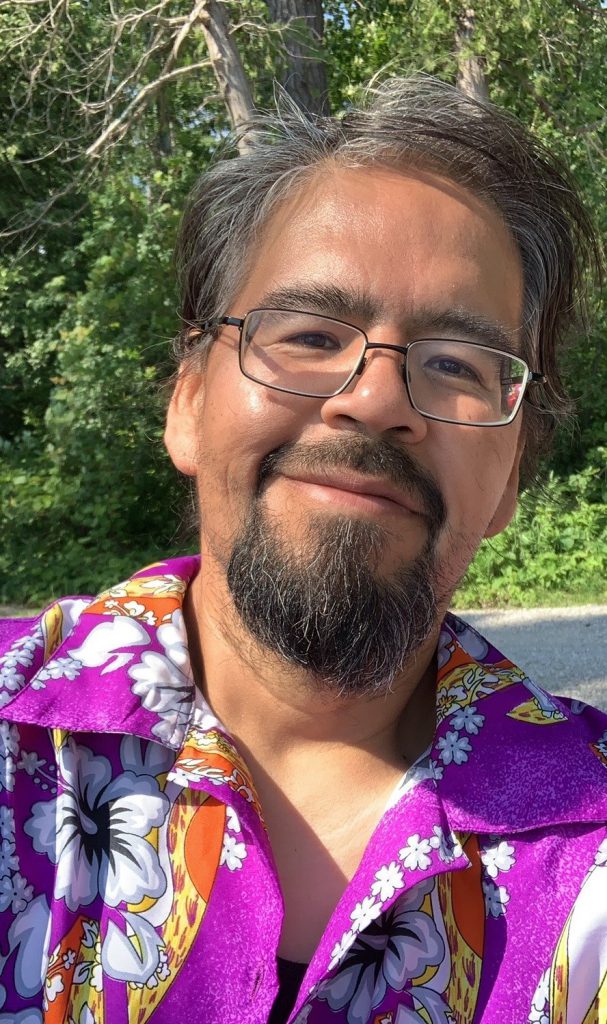 A person wearing a purple Hawaiian shirt smiles at the camera in a selfie while standing outside.