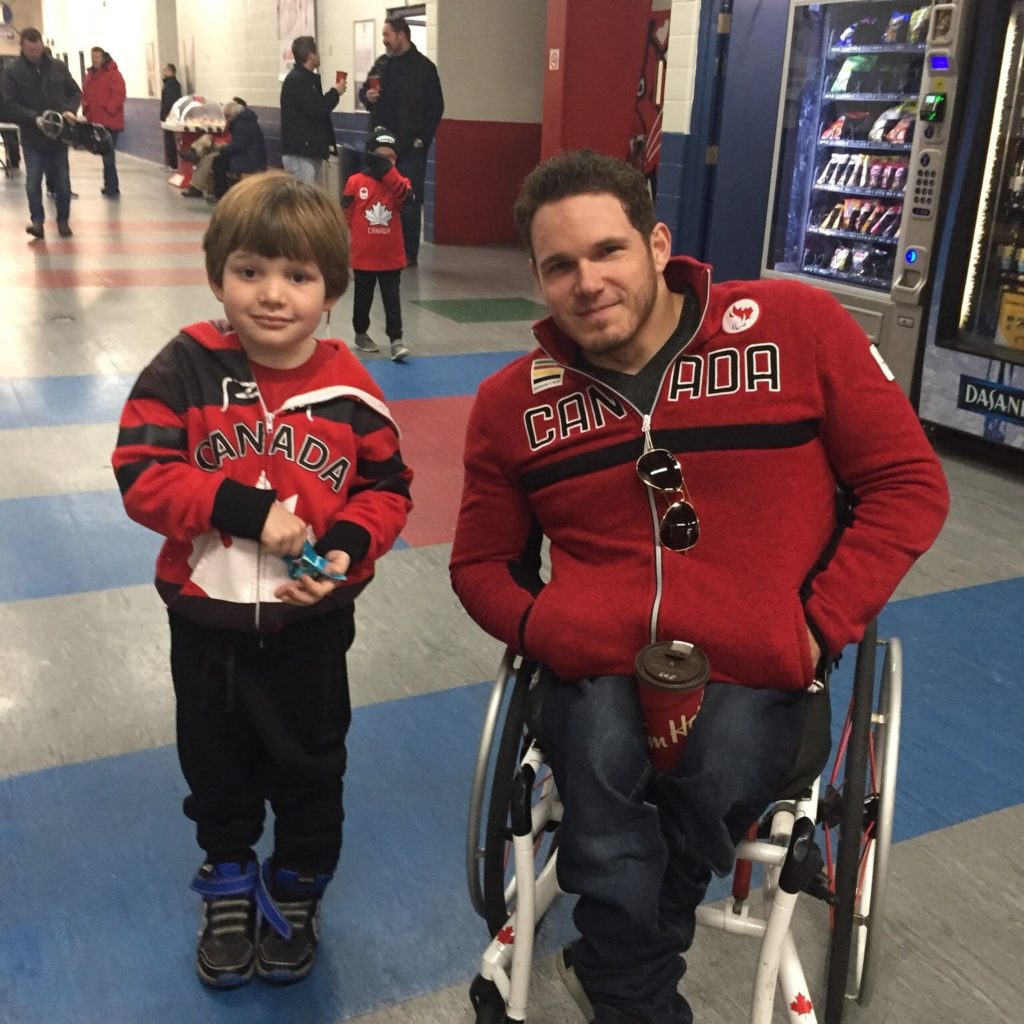 A person with short brown hair, red sweater reading "Canada," blue jeans and sitting in a wheelchair, smiles for a photo with a child who's also wearing a red sweater reading "Canada." 