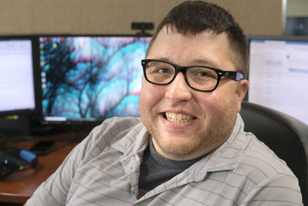 A smiling male with short dark hair iand glasses. He's sitting in front of a computer and is wearing a grey striped shirt.