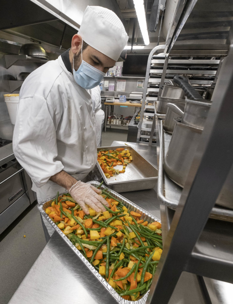 A young male wearing a white chef uniform looking at a large pan of vegetables