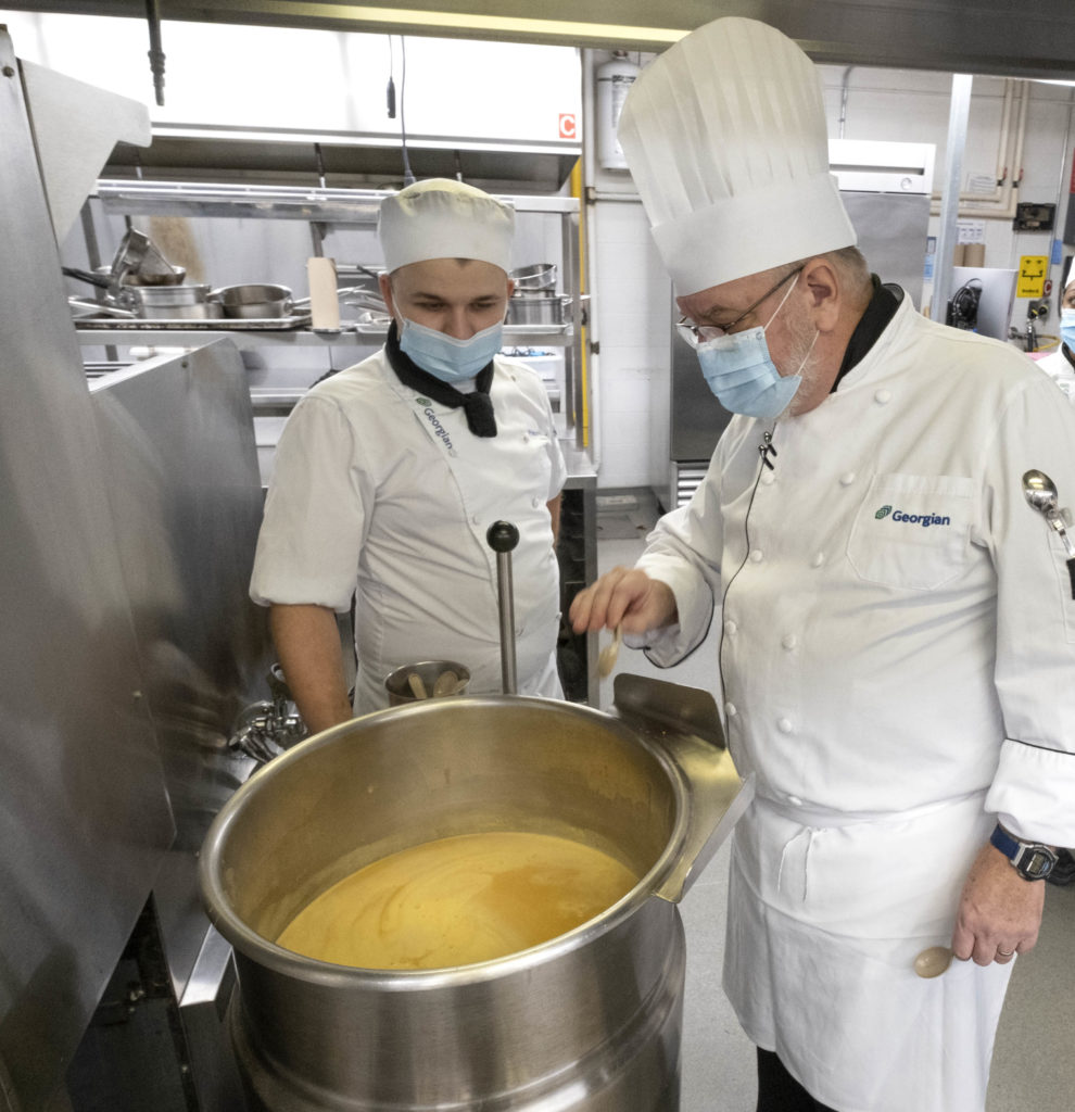 Two males in white chef uniforms looking at a pot of gravy in a kitchen