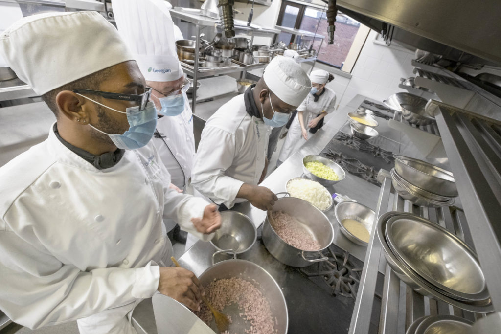 A line of people wearing white chef uniforms in a kitchen stirring pots on a stove.