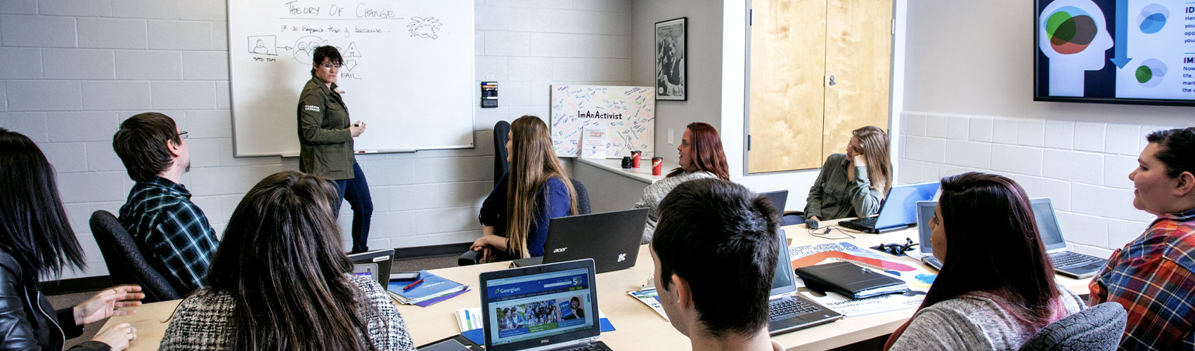 a group of students sitting at a table with laptops as a speaker makes notes on a whiteboard