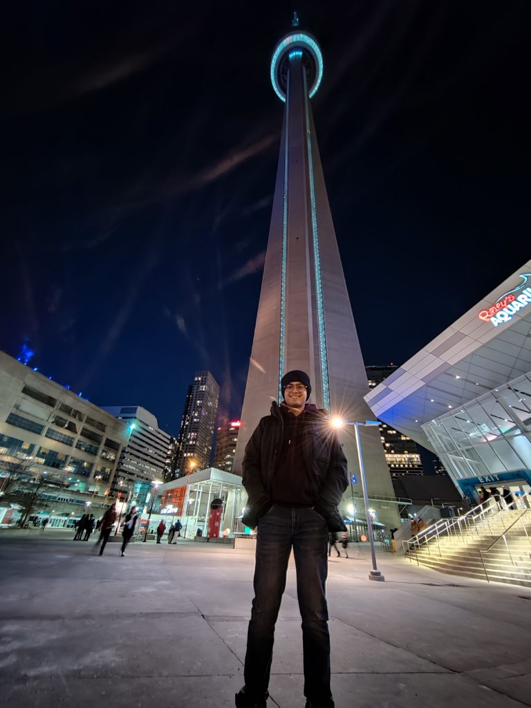 A person dressed in black coat, pants, hat and glasses, smiles for a photo in front of a tall tower.