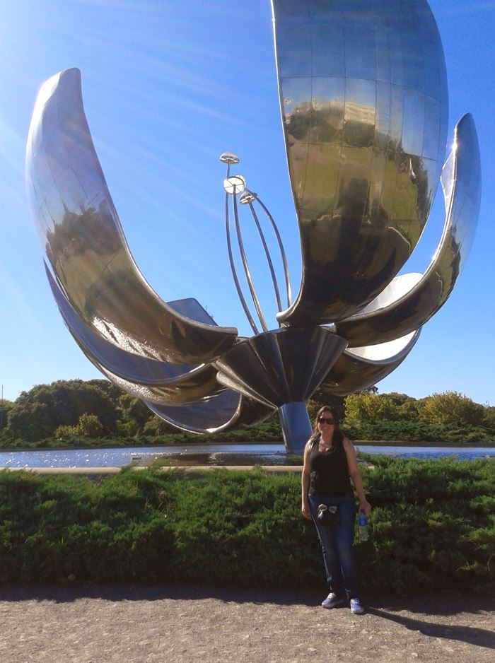 A person stands outside and smile in front of a large, silver metal statue of a flower.