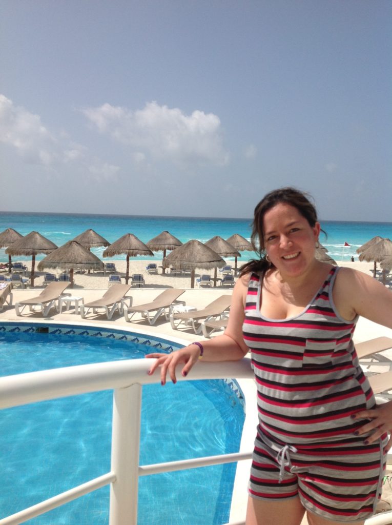 A person smiles and leans against a railing in front of a pool with an ocean and lounge chairs in the background.