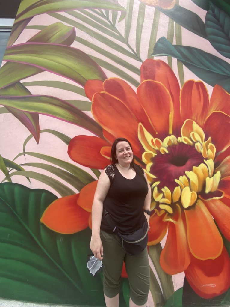 A person smiles in front of a large outdoor wall mural featuring a red and yellow flower.