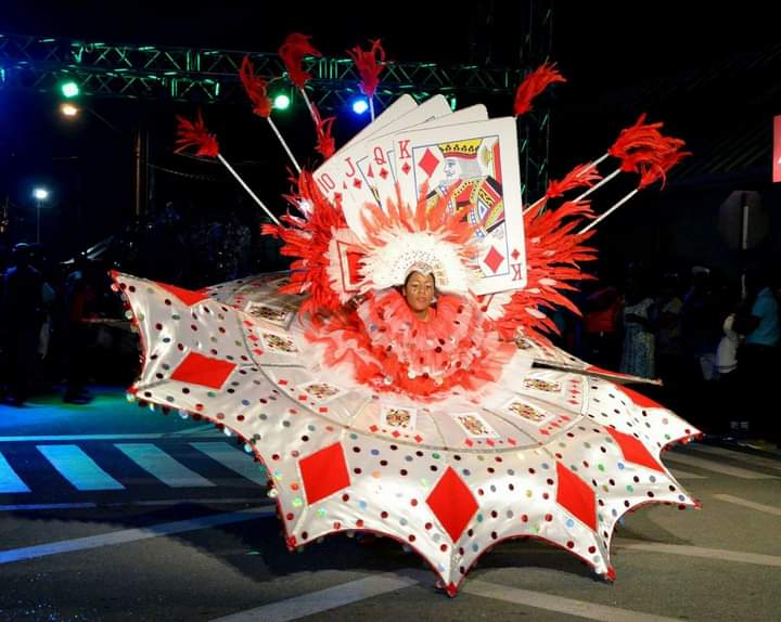 A person performs during a festival while dressed in a red and white outfit with oversized playing cards, tall feathers, and a sprawling skirt with diamonds and playing cards on it.
