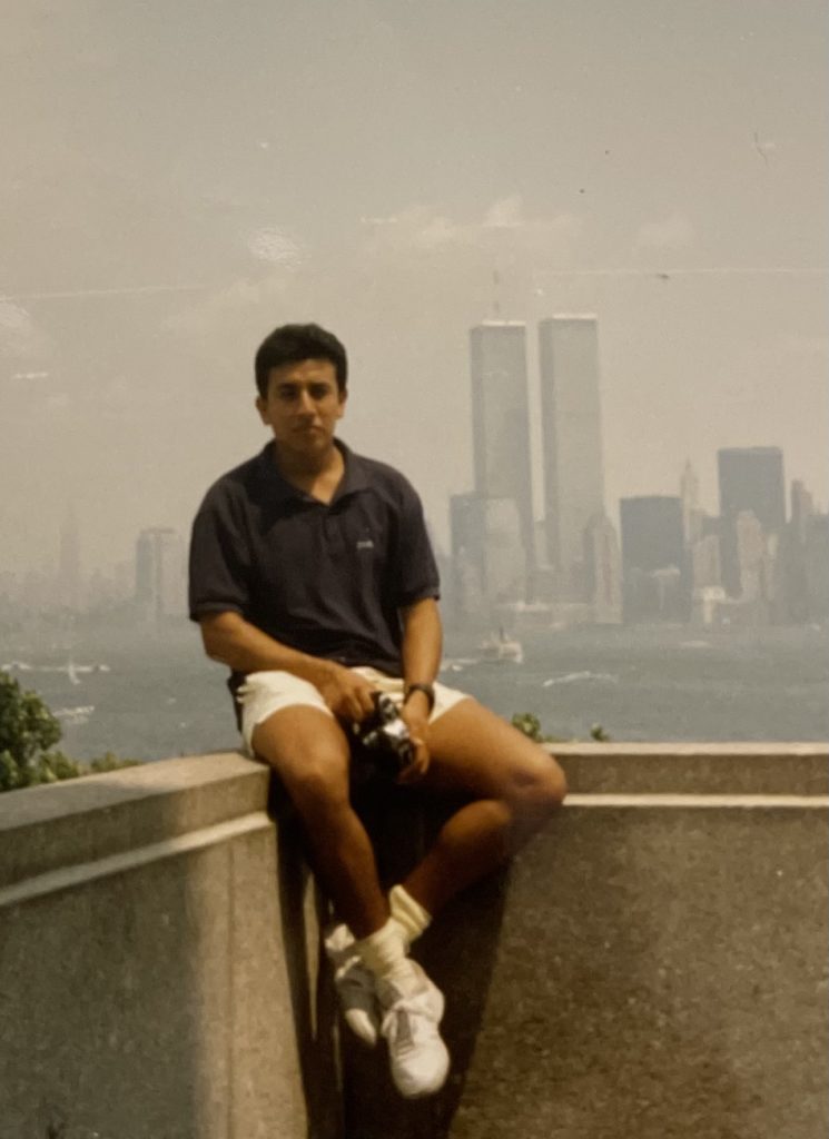 A person dressed casually sits on the edge of a wall with a river and the New York City skyline in the distance, showing the now-fallen Twin Towers.