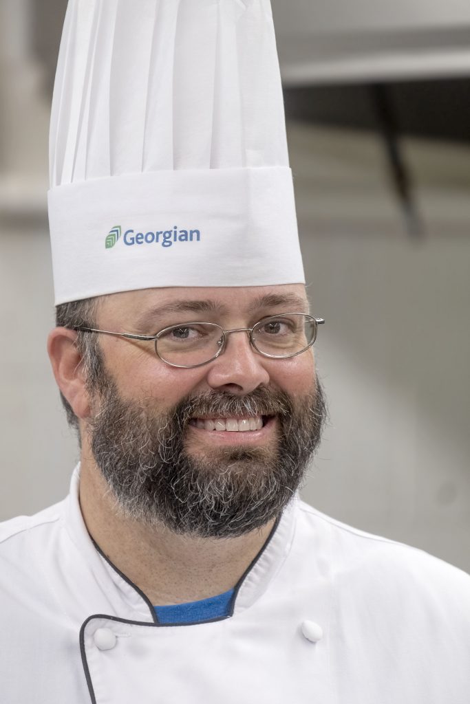 A smiling man with a beard wearing glasses, a white chef's hat and uniform