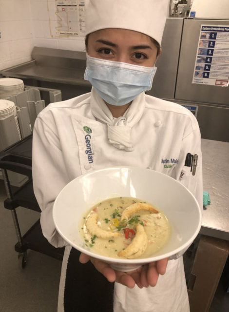 A young female wearing a white chef hat and uniform holding up a white bowl with food in it