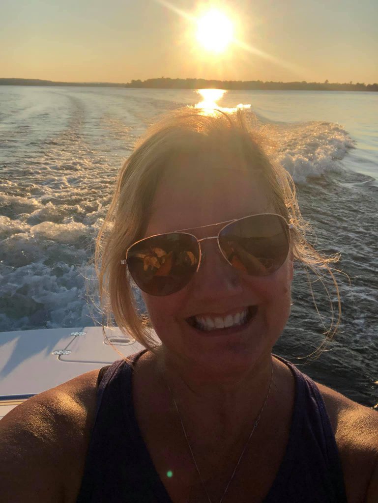 A headshot of a person with blonde hair, who's wearing sunglasses and a purple tank top, smiling while riding on the back of a boat, with waves churning behind and the sun shining over the horizon.