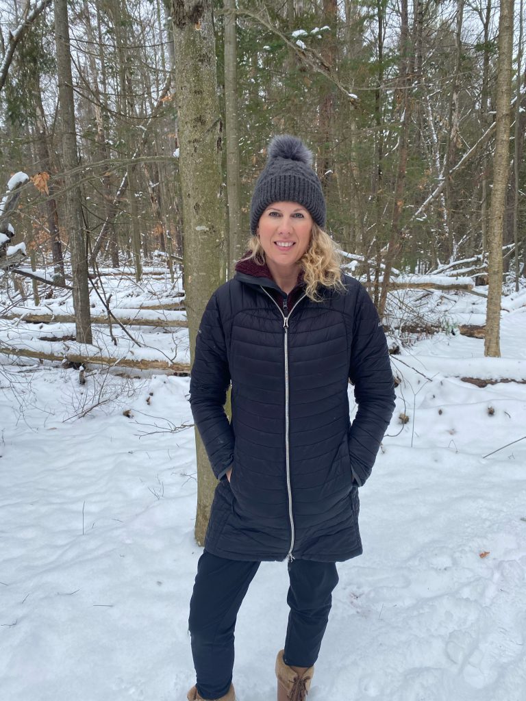 Person smiles at the camera while standing in a snowy forest.