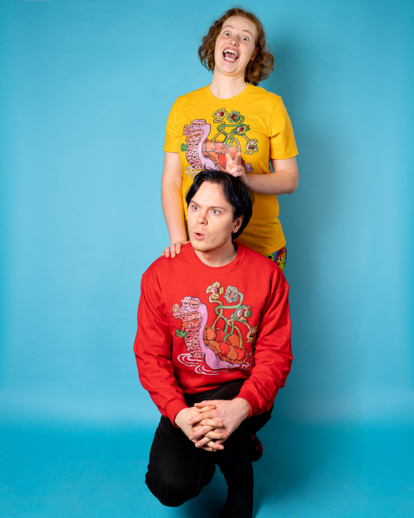One person with black hair, red sweatshirt and black pants, kneels in front of another person with curly, red hair, yellow shirt and pants, in front of a blue backdrop.