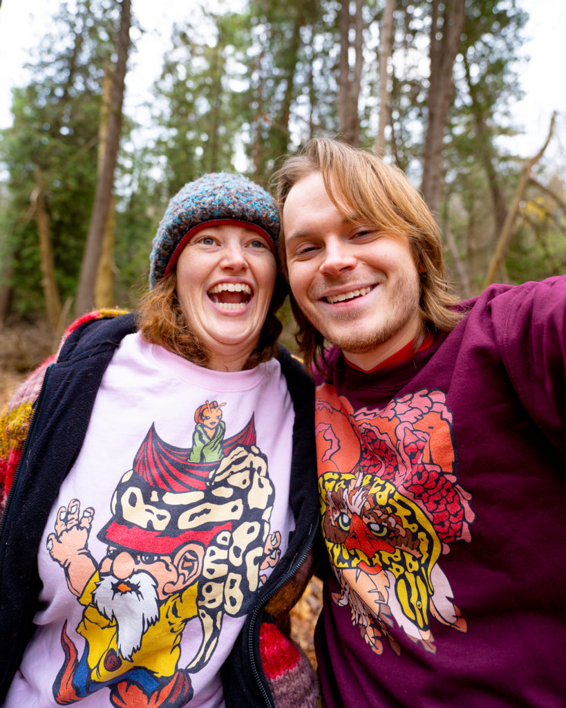 Two people stand outside in the woods and take a selfie. One is wearing a toque, colourful sweater, and a pink T-shirt with a colourful design on it. The other person has chin-length brown hair, beard, and is wearing a purple sweater with a colourful design on it.