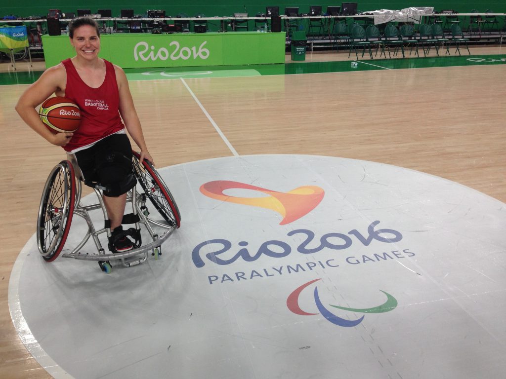 A person with brown hair and one leg, wearing a red tank top and blank shorts and shoes, holds a basketball and sits in a wheelchair on a gym floor that reads "Rio 2016 Paralympic Games."