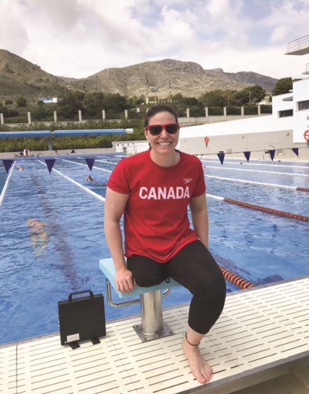 A person with brown hair and one leg, wearing a red shirt and sunglasses and black leggings, sits on a stool at the edge of a pool near a mountain range in the background.