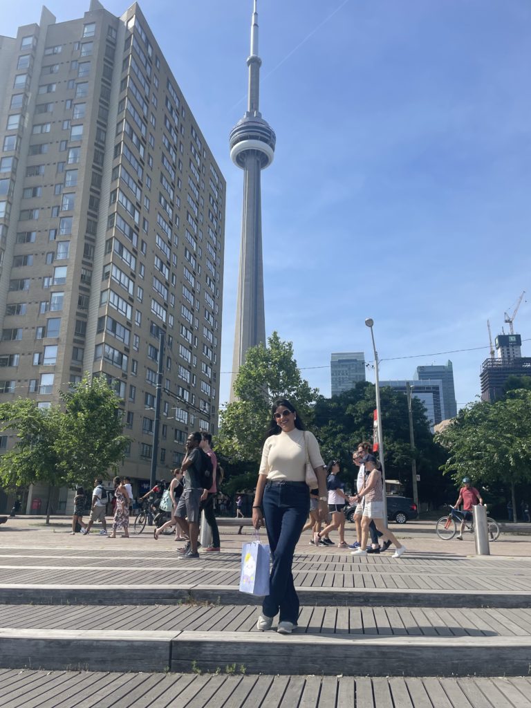 A person with long, black hair, sunglasses and dressed casually, holds a shopping bag and stands on a boardwalk, with tall buildings and the CN Tower behind them.