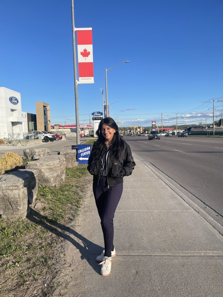 A person with long, black hair and dressed casually, stands on a city sidewalk with a Canada flag in the background and smiles at the camera.