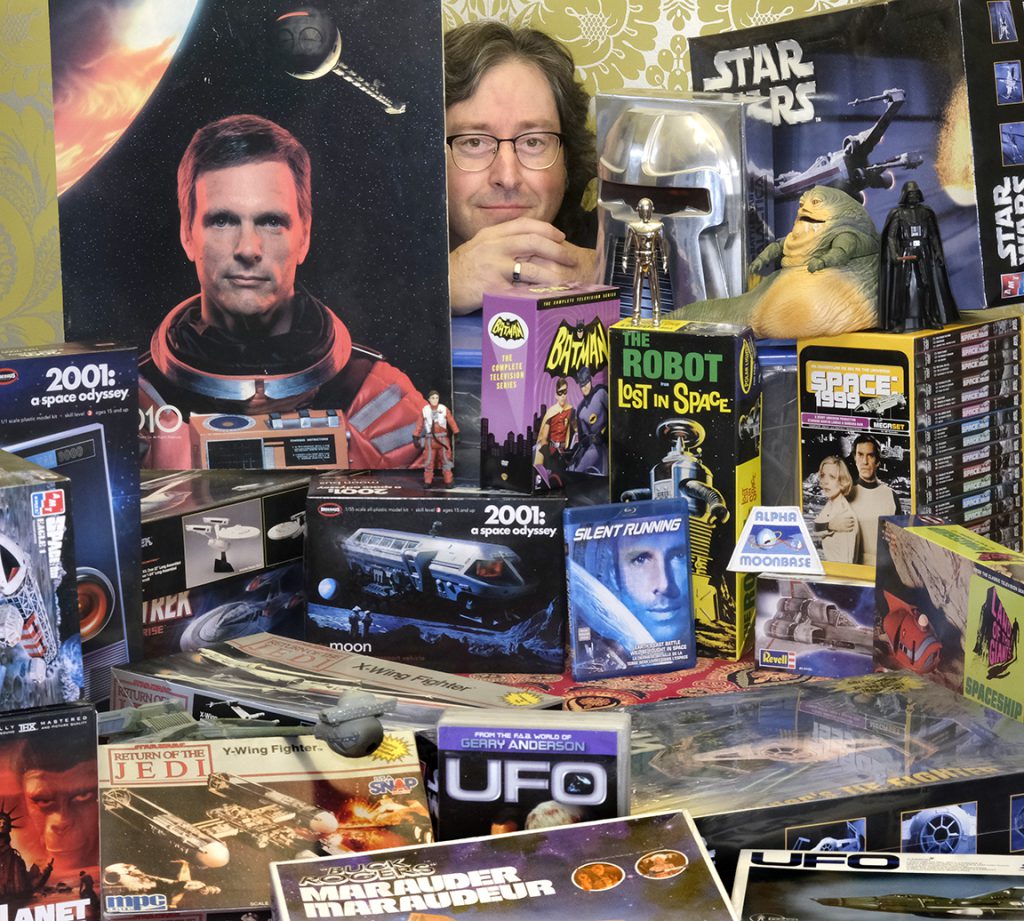 A person smiles as they peek out from behind a collection of science fiction paraphernalia.