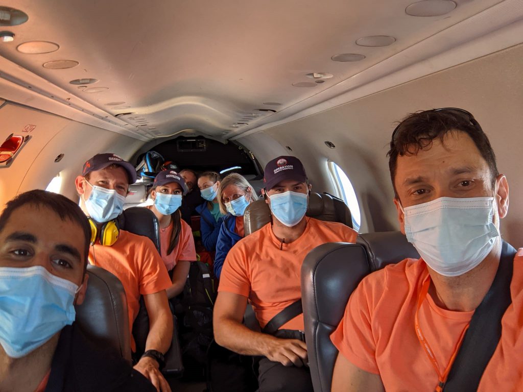 Eight people wearing orange shirts and blue face masks sit in chairs in a small airplane.