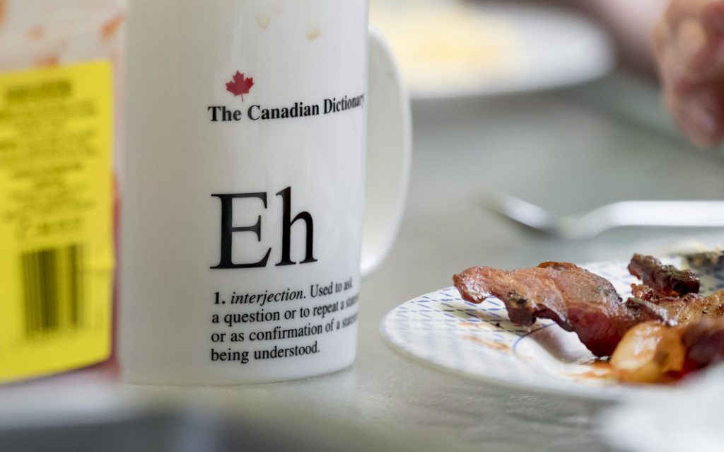 A mug that says "eh," sitting on the table next to some bacon