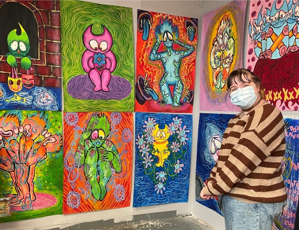 A person with brown hair pulled back, bangs, glasses, brown striped sweater and blue jeans, looks at the camera and stands in front of a wall of 10 colourful paintings.