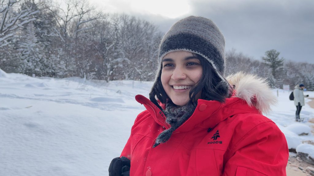 A person with short, brown hair and who's wearing a grey toque, red winter coat and a nose piercing, smiles while standing outside in the snow.
