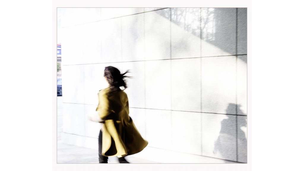 Blurry photo of a woman walking down a street in a mustard coloured coat