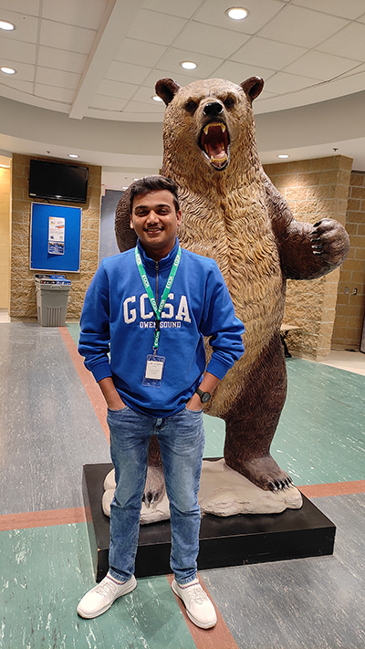 Hemchandran wearing a GCSA sweatshirt and badge, standing in front of the grizzly sculpture at the barrie campus