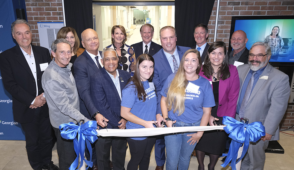 Fourteen people stand in three rows behind a ribbon on stanchions, and cut the ribbon to a new hallway wing.