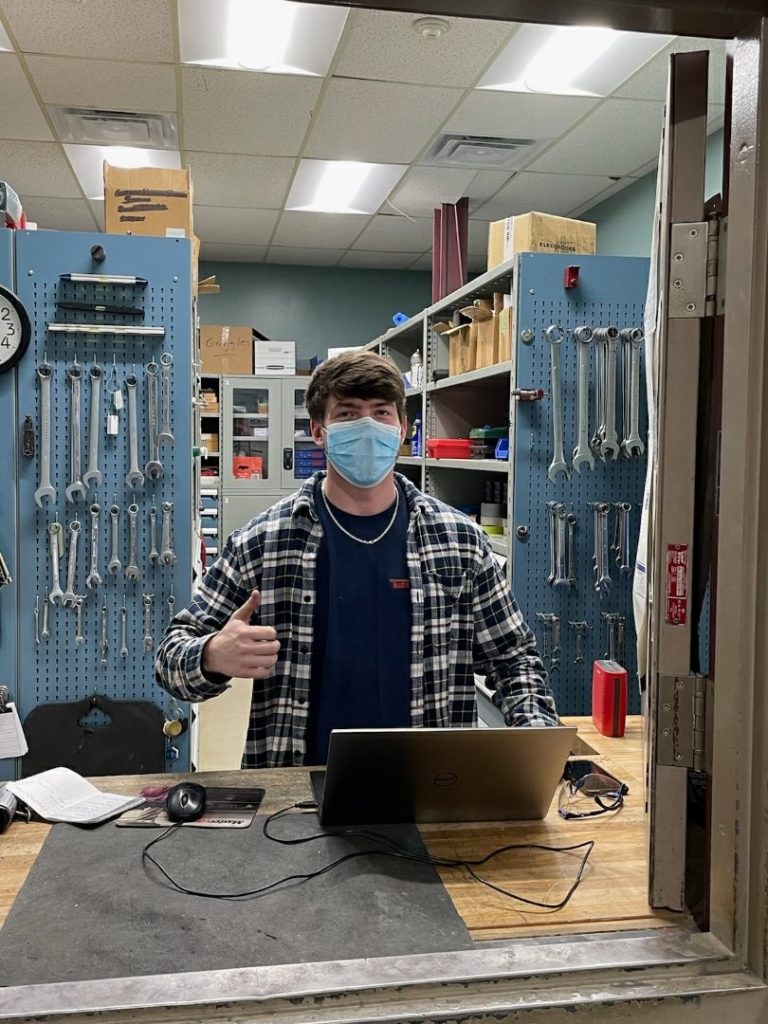 Ryan Miller standing behind the desk in the tool shop. He's got a mask on and is giving a thumbs up. Behind him you can see a wall of wrenches and cabinets.