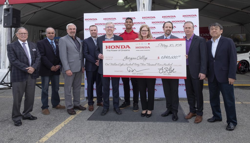A group of men and women in business attire standing in front of a tent opening. They're holding up a large cheque from Honda in the amount of $1,843,500.