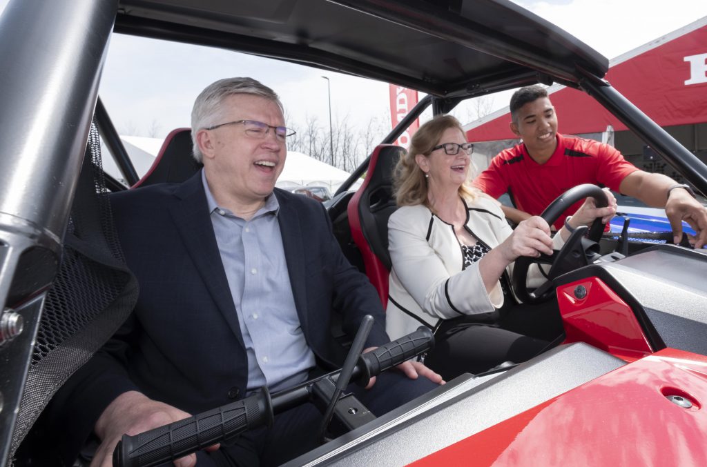 A gray-haired man with glasses and a business suit and a woman with blondish hair and glasses siting in a 4 x 4 vehicle. A young man in a red golf shirt is standing beside them. Everyone is smiling.
