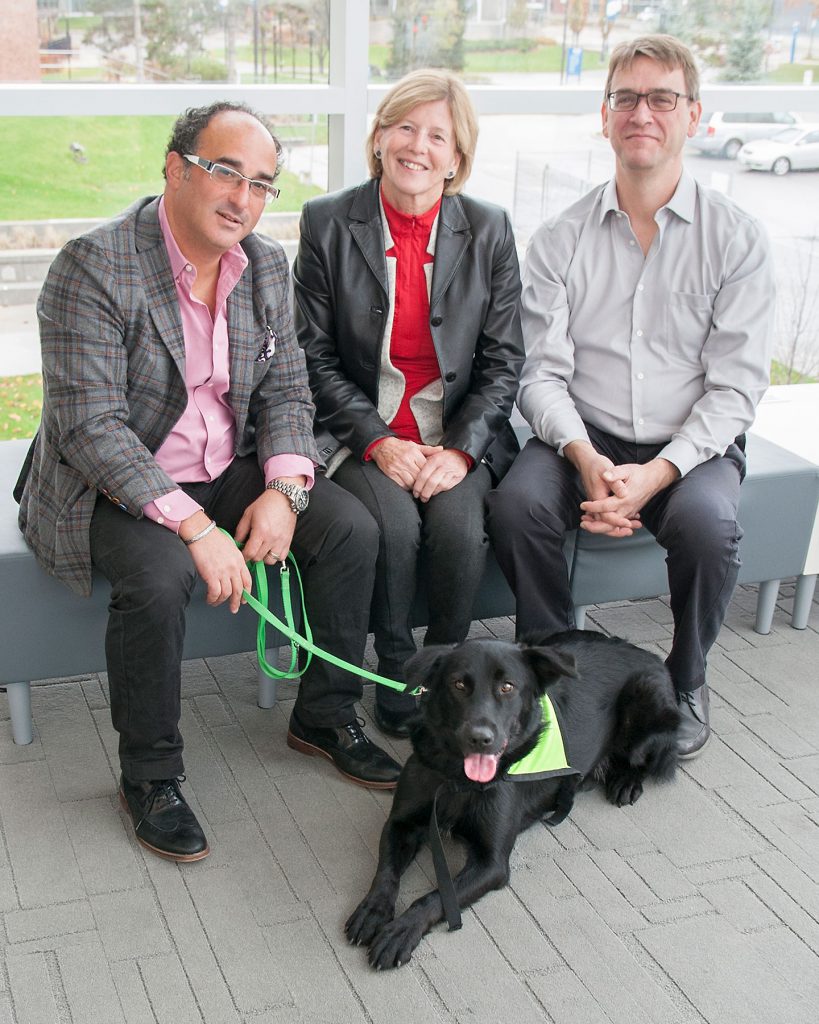 Three people sitting on a bench with a black dog. There are two older men flanked by an older blonde woman.