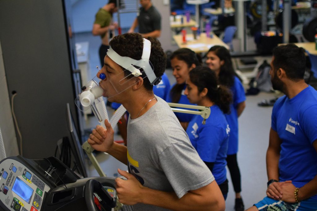 A young male jogging on a treadmill wearing a ventilator. There are young people wearing blue Georgian t-shirts standing in the room.