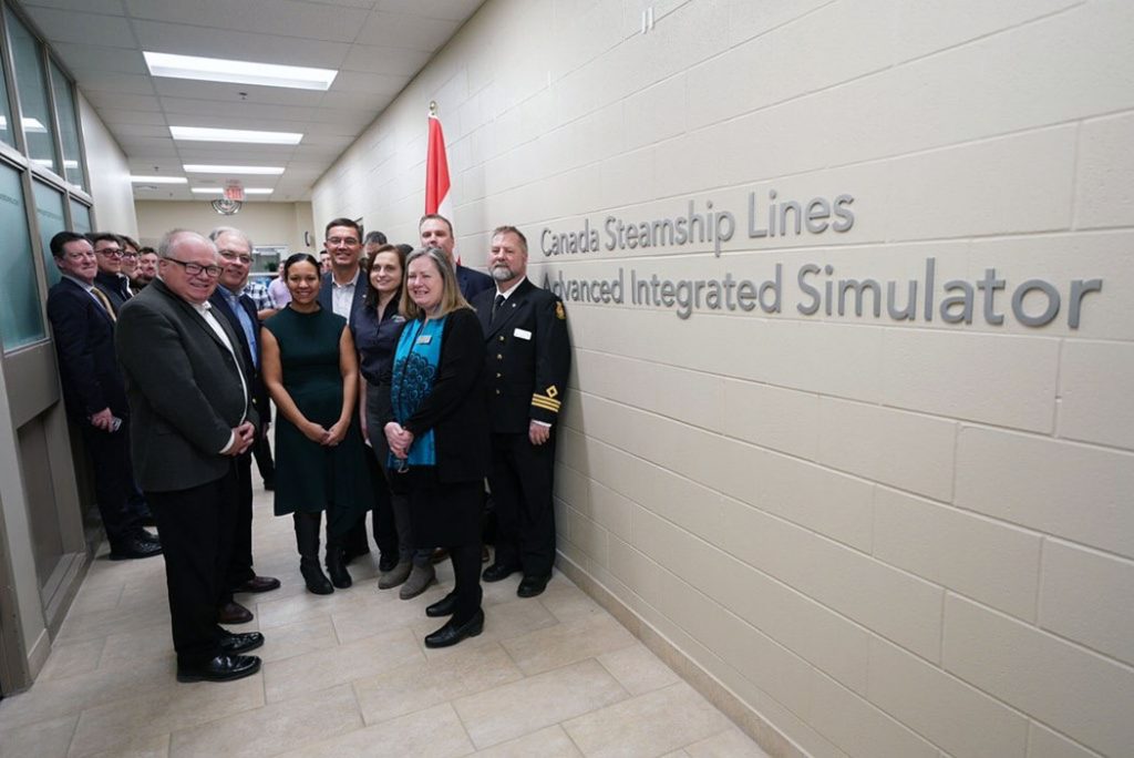 A group of people in business attire posed in a hallway with lettering on the wall reading: Canadian Steamship Lines Advanced Integrated Simulator