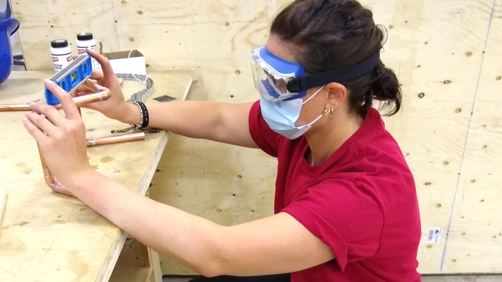 A female with dark hair wearing safety goggles, face mask and a red shirt standing in a wood shop measuring something