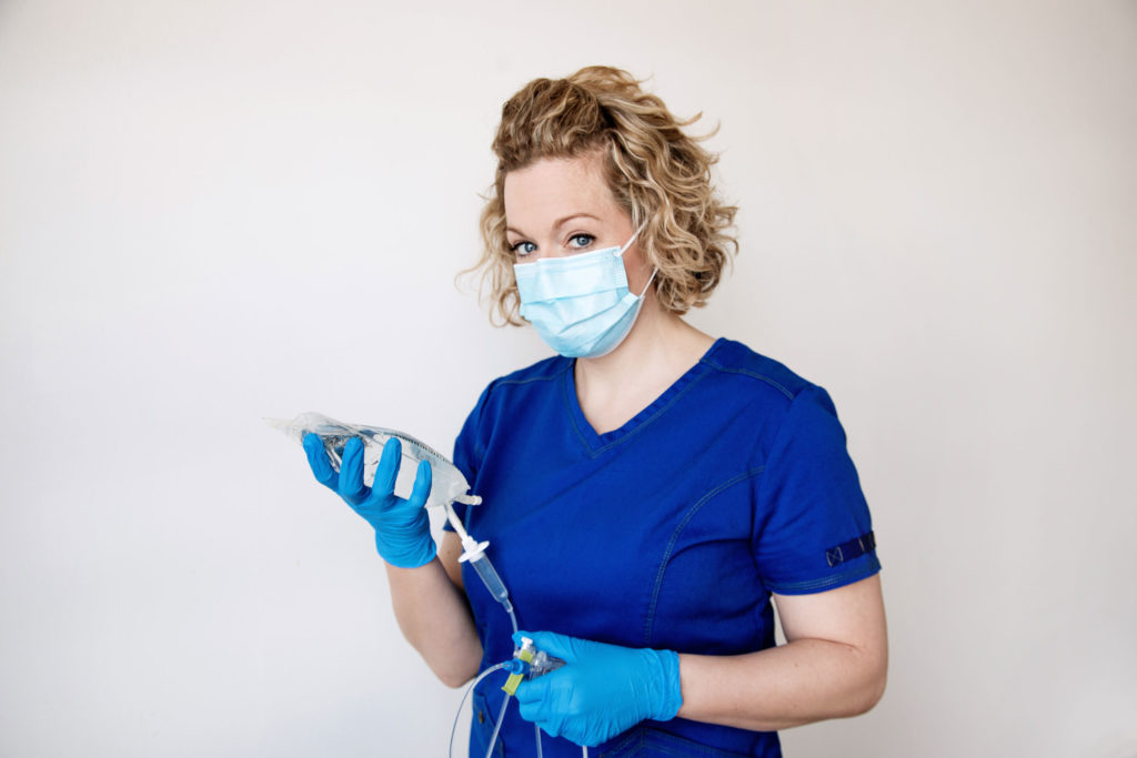 A blonde female wearing blue nursing uniform and a face mask. She's holding an IV bag